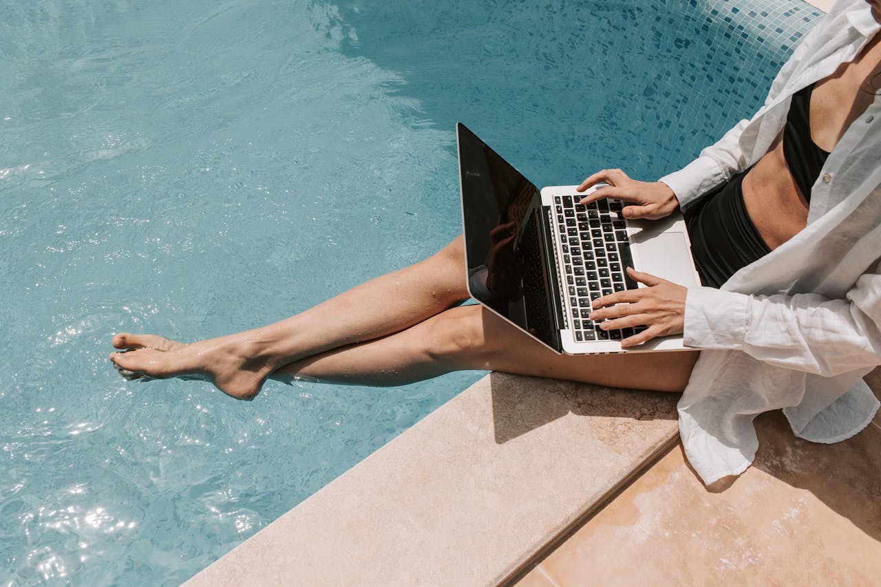 about-us-02 A woman in swimwear working on a laptop by the pool, showcasing summer remote work lifestyle.